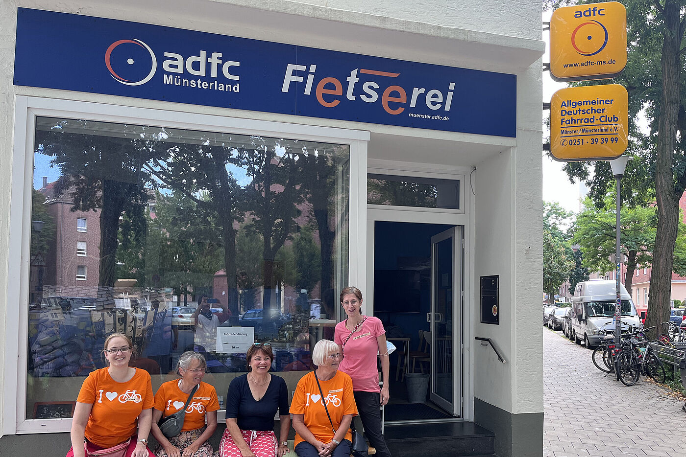 Fünf Frauen in orangener Fahrradkleidung vor der Geschäftsstelle des ADFC Münsterland an der Schaufensterscheibe mit blau weiß orangenem Schriftzug ADFC Münsterland Fietserei