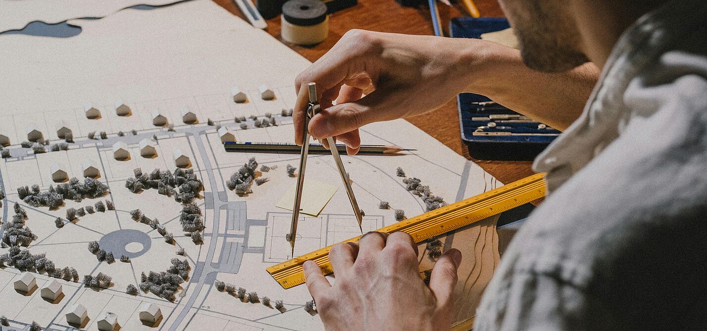 Ein Stadtplaner bei der Arbeit Ein Mann im Hemd ist von hinten zu sehen und zeichnet mit Lineal und Zirkel etwas auf einen Stadtplan.