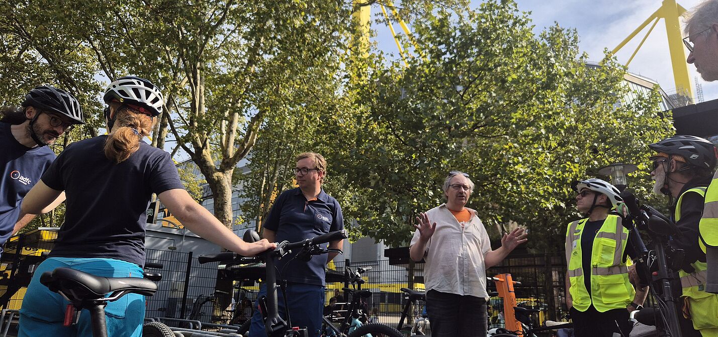 Menschen in Areal der Radwache, im Hintergrund das Stadion. 