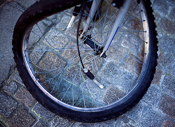 Nahaufnahme eines Fahrradvorderrads auf Kopfsteinpflaster. Die Felge ist verbogen und mehrere Speichen sind gebrochen oder hängen lose herab. Das Rad steht schief auf dem grauen Pflaster und ist offensichtlich nach einem Unfall nicht mehr fahrbereit.