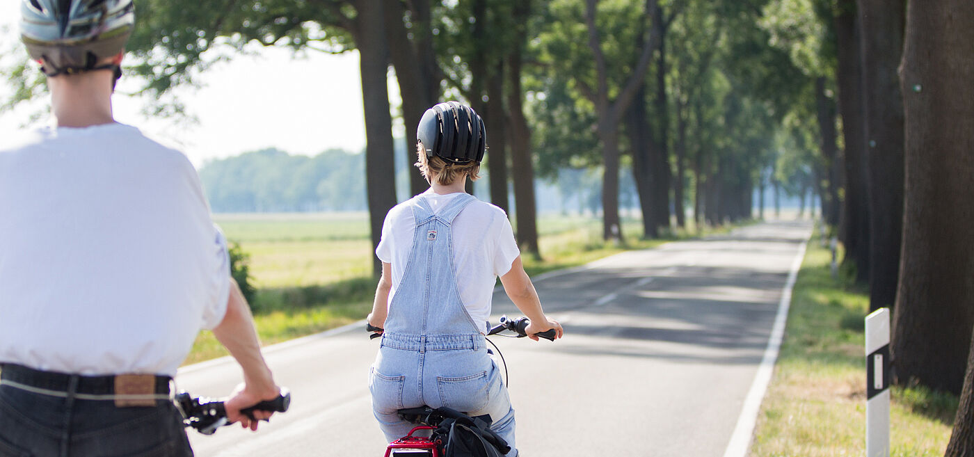 Zwei Personen mit Fahrradhelmen fahren auf einer schmalen Landstraße ohne separaten Radweg.
